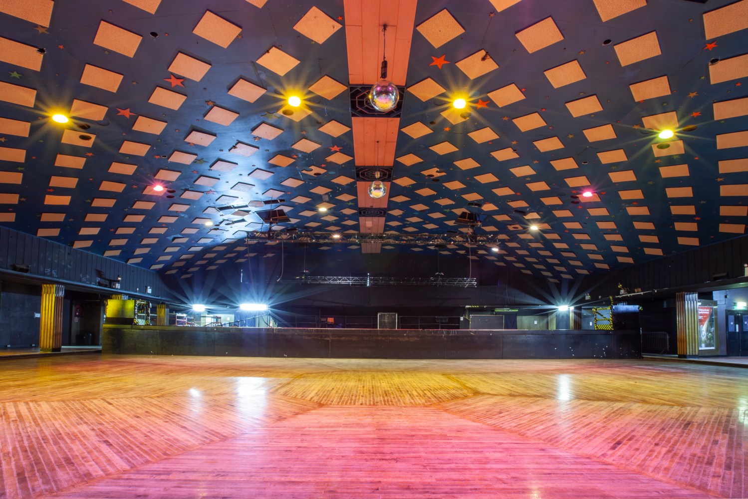 Empty venue with wooden floor, checkered blue ceiling, stage lights, and disco ball.